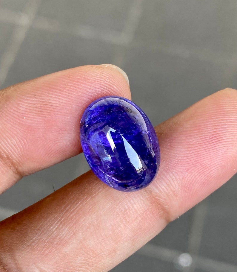 May include: A deep blue oval-shaped gemstone with a smooth, polished surface. The gemstone has a slight shimmer and is set in a clear, transparent setting.