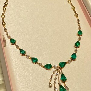 May include: A gold necklace featuring pear-shaped emeralds and small diamonds. The necklace has a delicate chain and a cluster of emeralds and diamonds hanging from the center. The necklace is displayed on a pink surface.