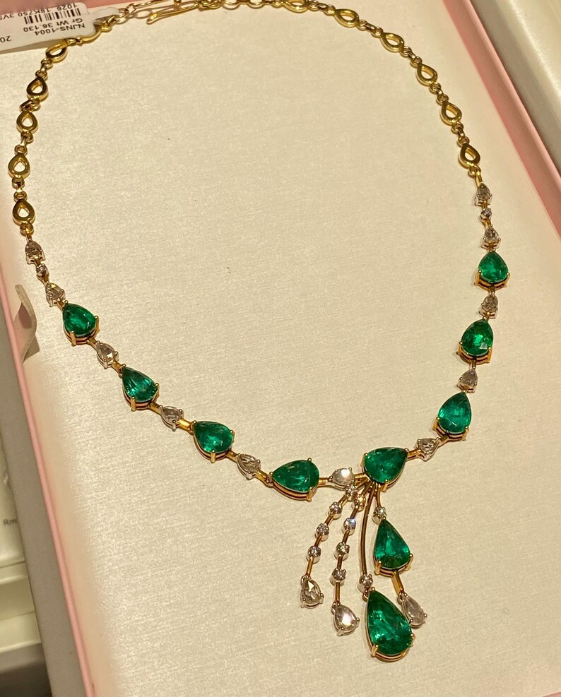 May include: A gold necklace featuring pear-shaped emeralds and small diamonds. The necklace has a delicate chain and a cluster of emeralds and diamonds hanging from the center. The necklace is displayed on a pink surface.