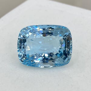 May include: A cushion-cut aquamarine gemstone with a light blue color. The gemstone is faceted and has a high degree of brilliance.