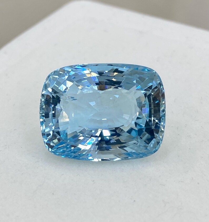 May include: A cushion-cut aquamarine gemstone with a light blue color. The gemstone is faceted and has a high degree of brilliance.