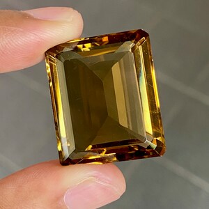 May include: A large, rectangular, golden-brown gemstone with a faceted cut. The gemstone is held between two fingers.