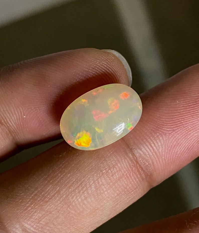 Pu&ograve; includere: Una pietra preziosa di opale ovale bianca con un arcobaleno di colori, tra cui giallo, arancione, rosso, verde e blu.
