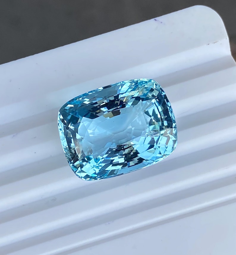 May include: A cushion-shaped aquamarine gemstone with a light blue color. The gemstone is faceted and has a high degree of brilliance.
