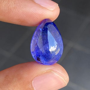 May include: A faceted, teardrop-shaped, blue gemstone. The gem is held between two fingers, showcasing its brilliant, transparent quality. The stone's facets reflect light, creating a sparkling effect. The color is a deep, rich blue.