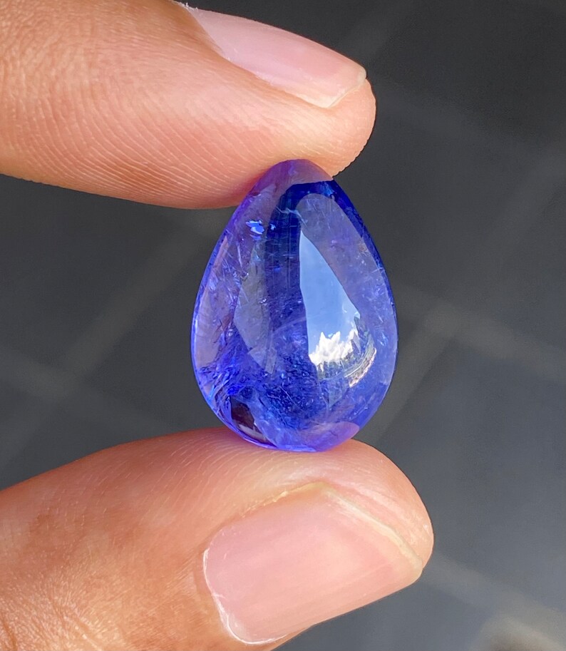 May include: A faceted, teardrop-shaped, blue gemstone. The gem is held between two fingers, showcasing its brilliant, transparent quality. The stone's facets reflect light, creating a sparkling effect. The color is a deep, rich blue.