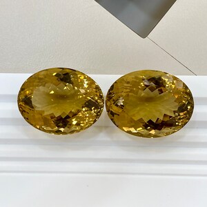 May include: Two oval-shaped, faceted, golden citrine gemstones on a white surface.