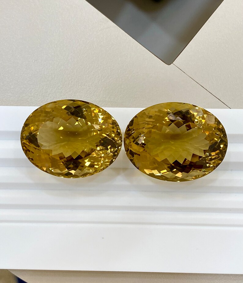 May include: Two oval-shaped, faceted, golden citrine gemstones on a white surface.