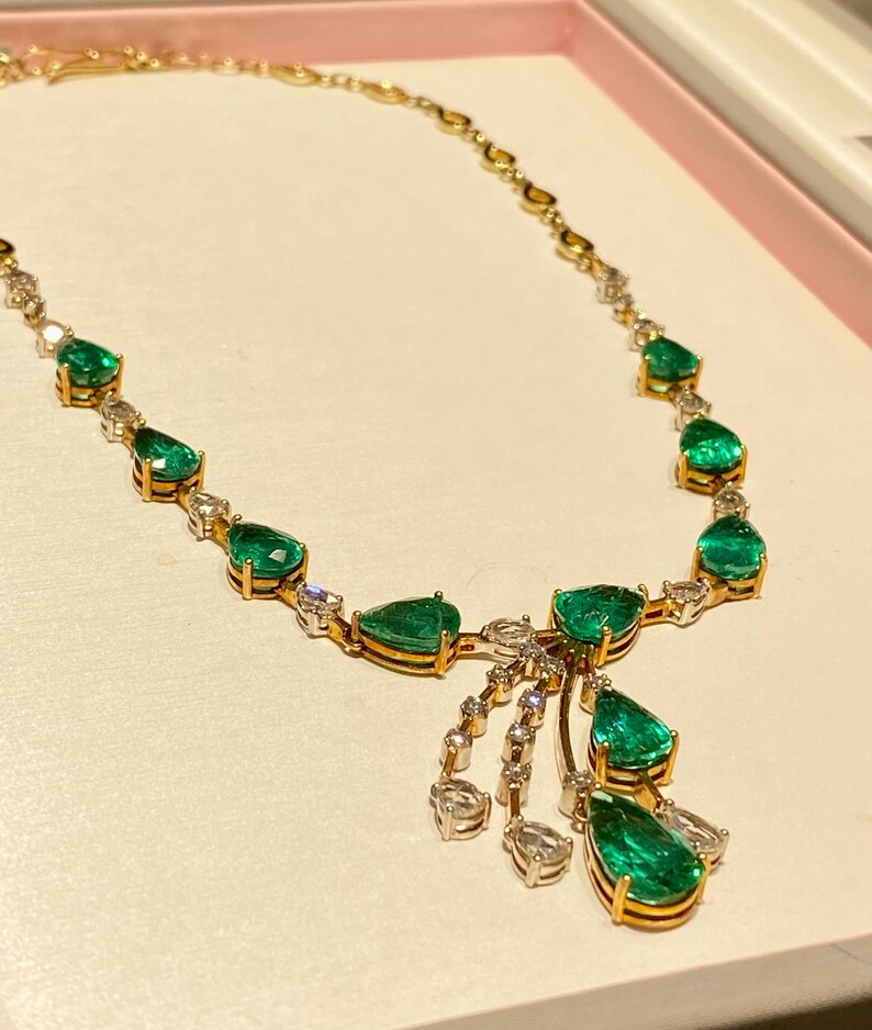 May include: A gold necklace featuring pear-shaped emeralds and small diamonds. The necklace has a delicate chain and a cluster of emeralds and diamonds that hang down. The emeralds are a vibrant green color.