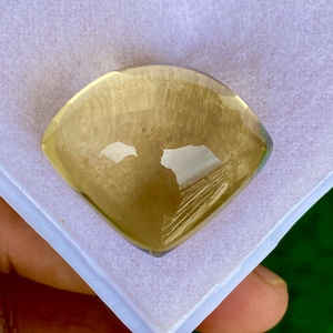 May include: A faceted, yellow-toned gemstone with a unique, shield-like shape. The gem is set against a white, textured background, and the light reflects off its polished surfaces. The gemstone is likely a citrine or similar quartz variety.