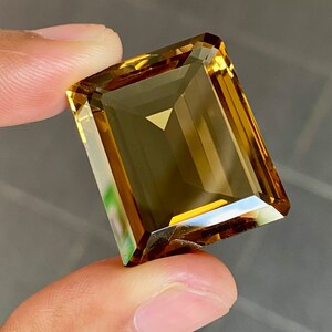 May include: A large, rectangular, golden-brown gemstone with a faceted cut. The gemstone is held in a hand.