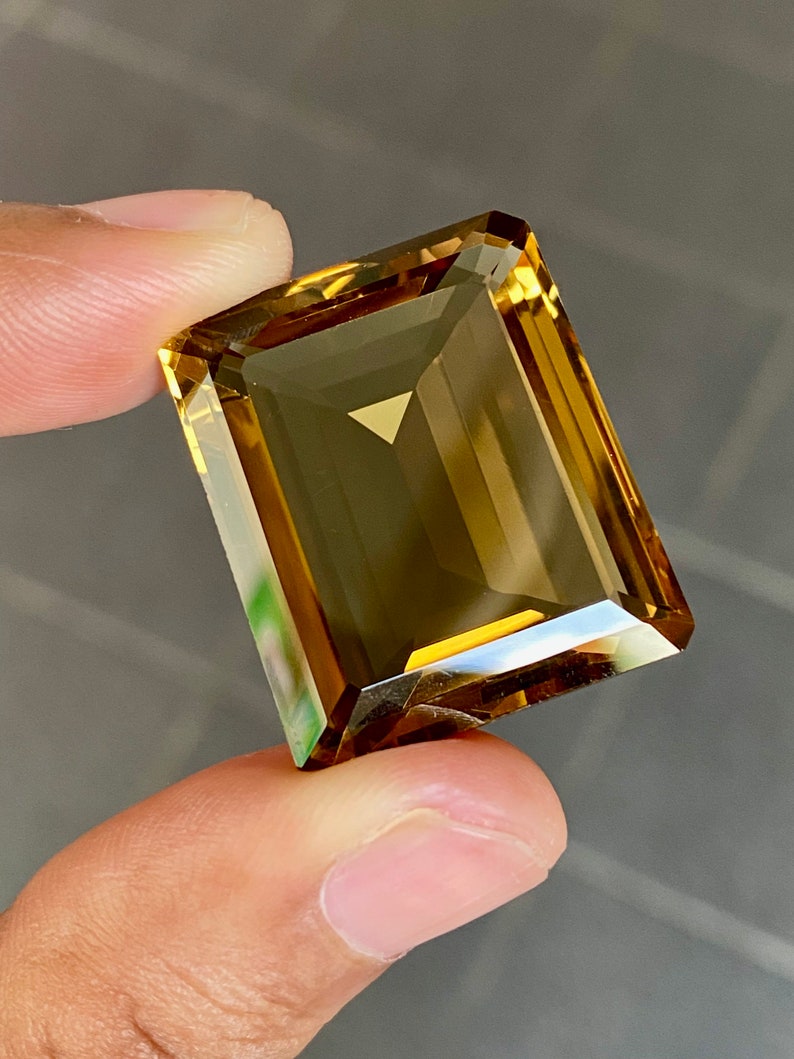 May include: A large, rectangular, golden-brown gemstone with a faceted cut. The gemstone is held in a hand.