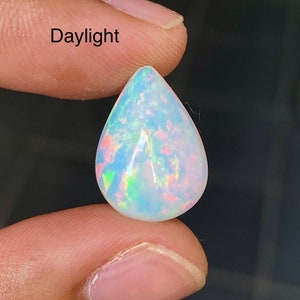 Può includere: Una pietra preziosa di opale a forma di lacrima con una base bianca e bagliori iridescenti di rosa, verde e blu. La pietra è tenuta da un dito, e la parola "Daylight" è visibile sullo sfondo.