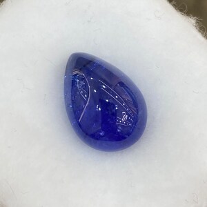 May include: A polished, teardrop-shaped gemstone in a deep, saturated blue hue. The stone has a smooth, reflective surface, with subtle internal patterns. The gem is set against a soft, white background, highlighting its color and form.