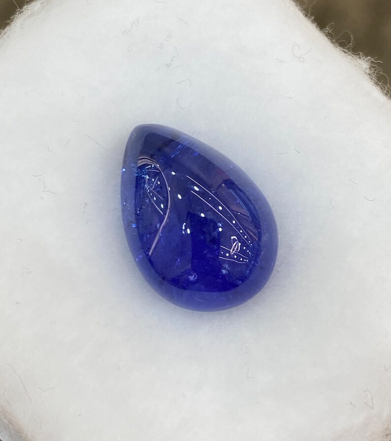 May include: A polished, teardrop-shaped gemstone in a deep, saturated blue hue. The stone has a smooth, reflective surface, with subtle internal patterns. The gem is set against a soft, white background, highlighting its color and form.