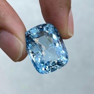 May include: A large, light blue aquamarine gemstone with a cushion cut. The gemstone is faceted and has a brilliant shine.