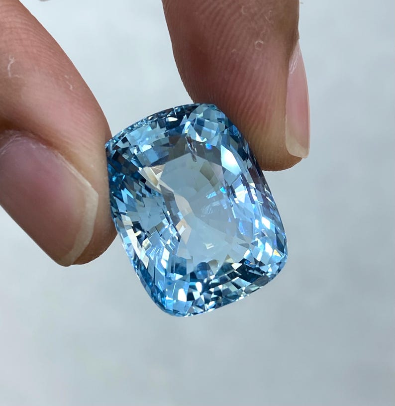May include: A large, light blue aquamarine gemstone with a cushion cut. The gemstone is faceted and has a brilliant shine.