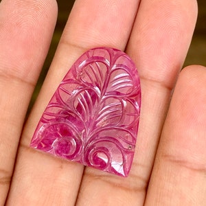 May include: A carved, ruby-colored gemstone with an arched top. The gemstone features an intricate floral design carved into its surface. The stone is held in a hand, showcasing its color and detail. The gemstone is a cabochon.