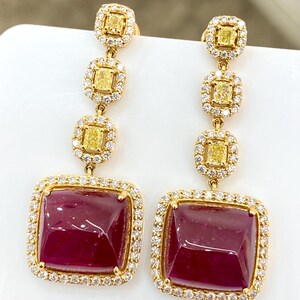 May include: A pair of gold drop earrings featuring three vertically aligned yellow square-cut stones, each surrounded by a halo of small white diamonds, ending in a large square-cut deep red gemstone framed by a border of white diamonds.