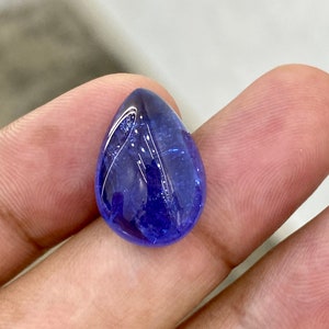 May include: A polished, teardrop-shaped gemstone with a deep blue hue. The gem has a smooth, reflective surface, with subtle internal patterns. The stone is held in a hand, showcasing its size and color. The gem is likely a tanzanite.