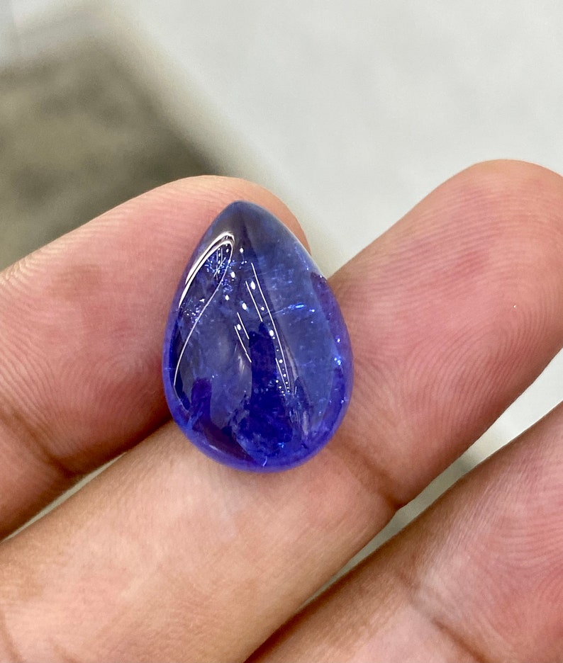 May include: A polished, teardrop-shaped gemstone with a deep blue hue. The gem has a smooth, reflective surface, with subtle internal patterns. The stone is held in a hand, showcasing its size and color. The gem is likely a tanzanite.