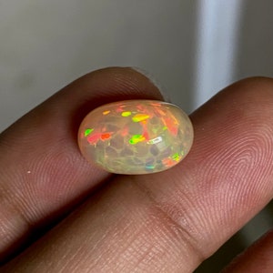 May include: A light brown oval opal gemstone with a white base and flashes of red, orange, yellow, and green.