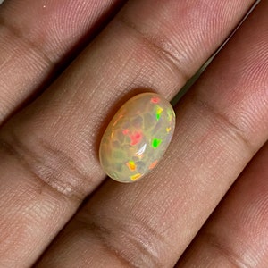 Pu&ograve; includere: Un cabochon di opale bianco con un bagliore verde chiaro, arancione e rosso. L'opale &egrave; di forma ovale e ha una superficie liscia e lucida.