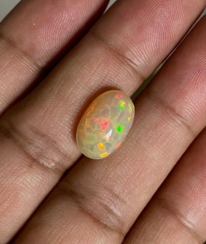 Pu&ograve; includere: Un cabochon di opale bianco con un bagliore verde chiaro, arancione e rosso. L'opale &egrave; di forma ovale e ha una superficie liscia e lucida.
