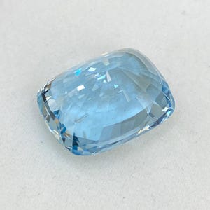 May include: A light blue aquamarine gemstone with a cushion cut. The gemstone is faceted and has a high degree of brilliance.