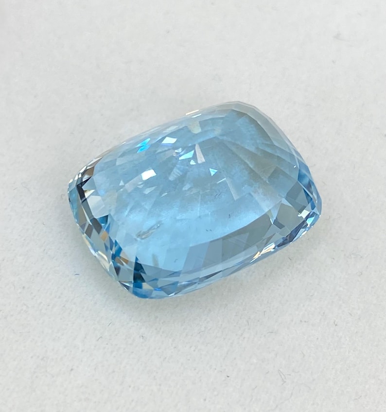 May include: A light blue aquamarine gemstone with a cushion cut. The gemstone is faceted and has a high degree of brilliance.