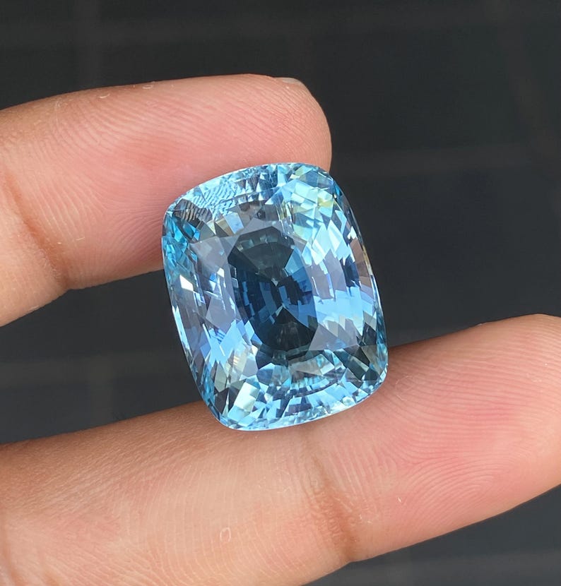 May include: A large, cushion-cut aquamarine gemstone with a light blue color. The gemstone is faceted and has a high degree of brilliance.