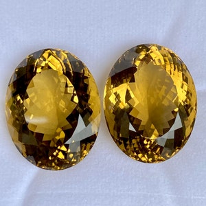 May include: Two oval-shaped, faceted gemstones with a golden yellow color. The gemstones have a slight brown hue and are set against a white background.