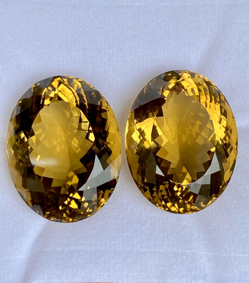 May include: Two oval-shaped, faceted gemstones with a golden yellow color. The gemstones have a slight brown hue and are set against a white background.