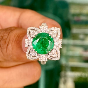 May include: A green emerald gemstone ring with a floral design set in white gold. The ring is surrounded by smaller diamonds.