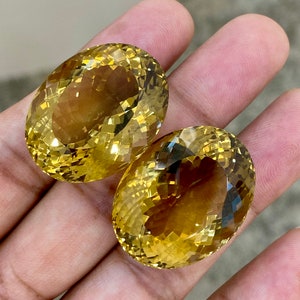 May include: Two oval-shaped, faceted, yellow citrine gemstones. The citrines are a light to medium yellow color and have a slight brown hue.