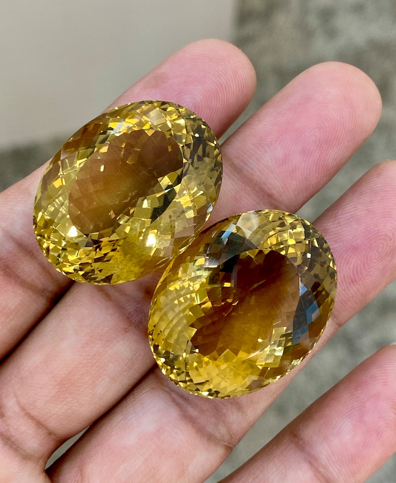 May include: Two oval-shaped, faceted, yellow citrine gemstones. The citrines are a light to medium yellow color and have a slight brown hue.