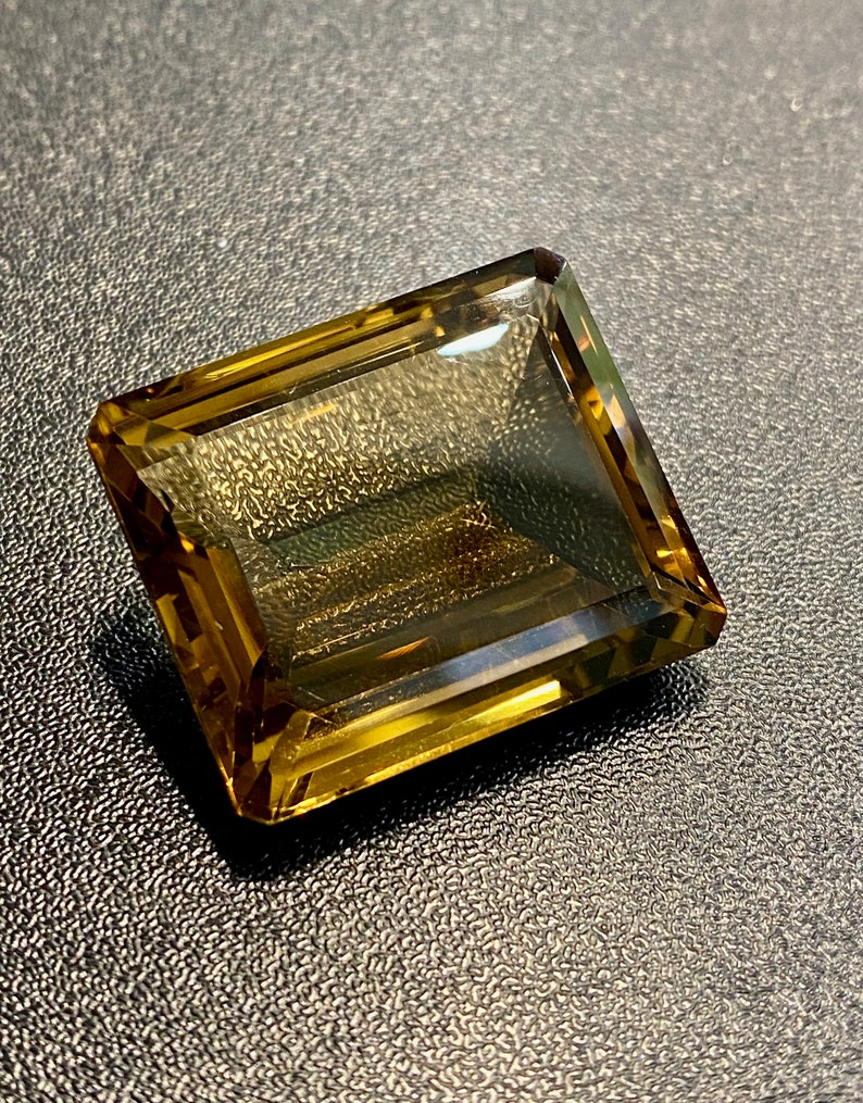 May include: A large, rectangular, faceted gemstone with a golden brown color. The gemstone is cut into a step cut style.