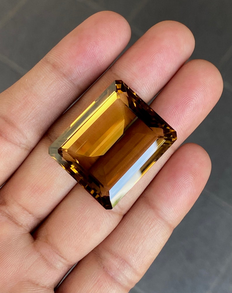 May include: A large, rectangular, faceted gemstone with a golden brown color. The gemstone is cut in an emerald shape.