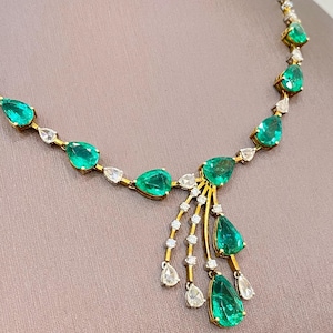 May include: A gold necklace with emerald green gemstones and clear, teardrop-shaped stones. The necklace has a delicate design with a central cluster of emeralds and dangling stones. The background is a neutral colour.