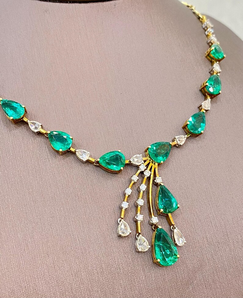 May include: A gold necklace featuring emerald-cut green gemstones and clear, teardrop-shaped stones. The necklace has a delicate design with a central cluster of emeralds and dangling stones, set against a neutral background.