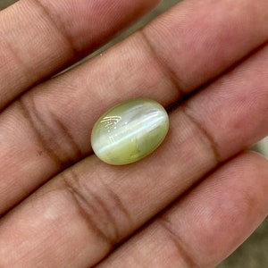 May include: A light green oval-shaped gemstone with a chatoyant effect. The stone is held in a hand.