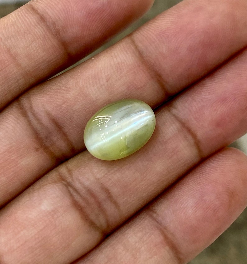 May include: A light green oval-shaped gemstone with a chatoyant effect. The stone is held in a hand.