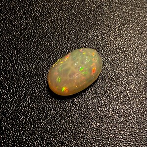 Pu&ograve; includere: Una pietra preziosa di opale bianco con un bagliore iridescente verde chiaro, rosa e arancio. La pietra preziosa di forma ovale &egrave; posizionata su uno sfondo nero.