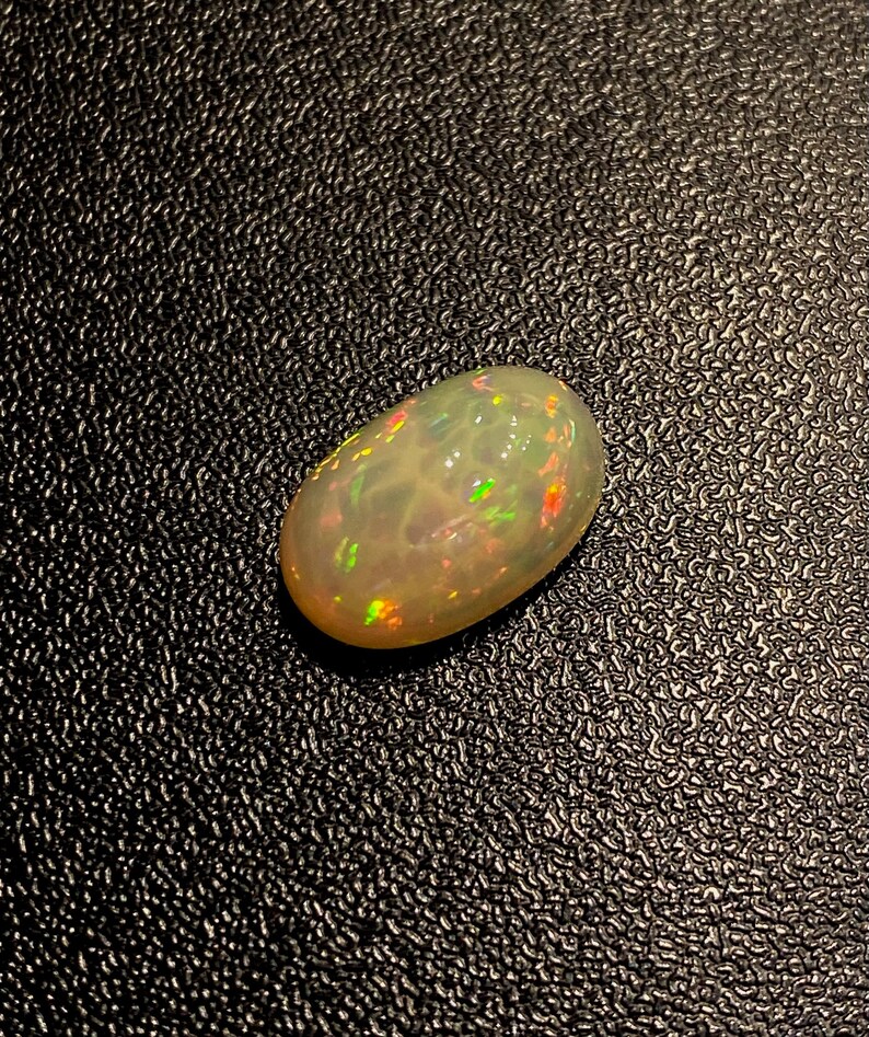 Pu&ograve; includere: Una pietra preziosa di opale bianco con un bagliore iridescente verde chiaro, rosa e arancio. La pietra preziosa di forma ovale &egrave; posizionata su uno sfondo nero.