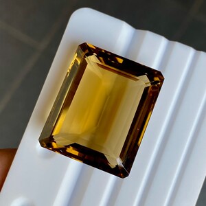 May include: A large, rectangular, golden brown gemstone with a faceted cut. The gemstone is displayed on a white surface.