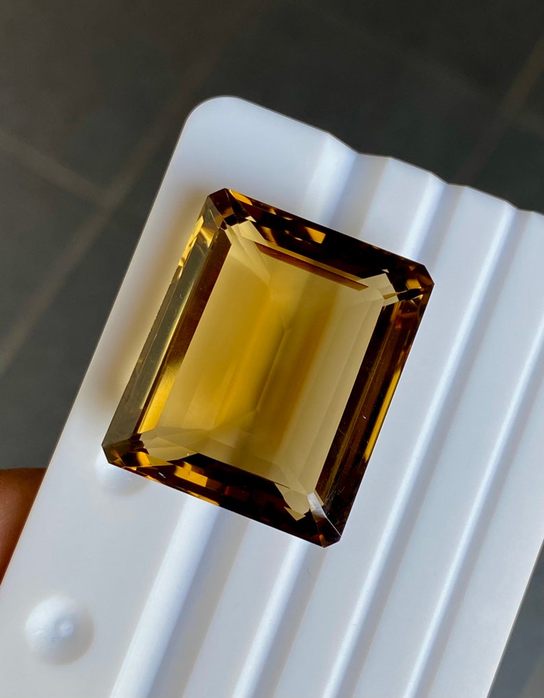 May include: A large, rectangular, golden brown gemstone with a faceted cut. The gemstone is displayed on a white surface.