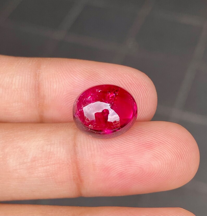 May include: A round, cabochon-cut, red gemstone with a high polish. The gemstone is set in a bezel setting and has a slight sparkle.