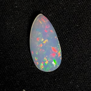 May include: A pear-shaped opal gemstone with a milky white base and vibrant flashes of red, green, and yellow. The gemstone is set against a black background, highlighting its iridescent qualities. The opal's shape is elongated and smooth.