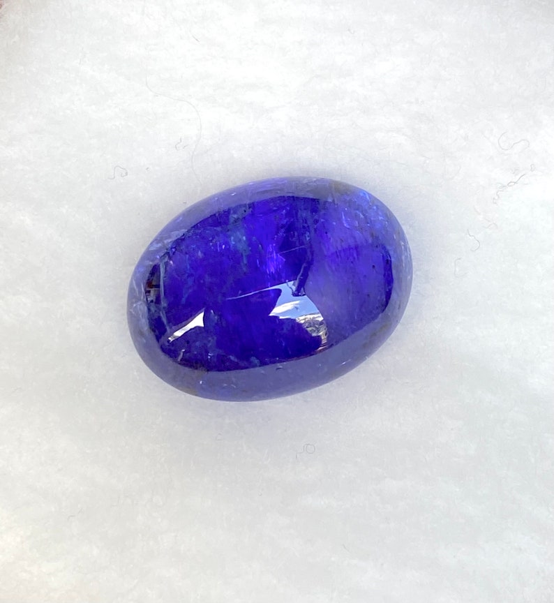 May include: A deep blue oval-shaped gemstone with a smooth, polished surface. The gemstone has a slight shimmer and is set against a white background.