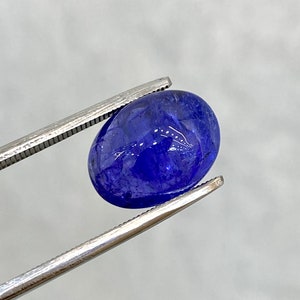 May include: A deep blue oval-shaped gemstone held by tweezers. The gemstone has a smooth, polished surface.